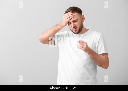 Junger Mann mit Glas Wasser, der an Kopfschmerzen auf hellem Hintergrund leidet Stockfoto