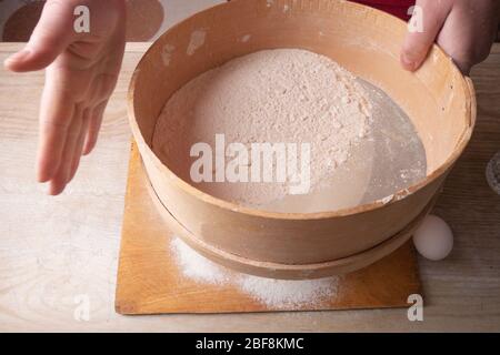 Große Schüssel, Schneidebrett, Sieb zum Sieben von Mehl auf dem Tisch. Weibliche Hände sieben Mehl in der Küche. Stockfoto