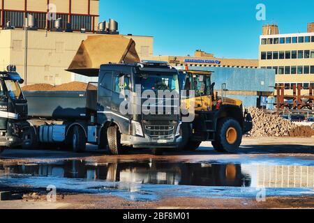 Komatsu Radlader Verladung Kies auf den Anhänger des schwarzen Volvo FH Kipper LKW auf der Baustelle an sonnigen Tag. Helsinki, Finnland. April 17, 2020 Stockfoto