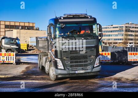 Schwarzer Volvo FH Kipper LKW mit einer Ladung Schotter verlässt Baustelle mit schweren Maschinen an sonnigen Tag. Helsinki, Finnland. 17. April 2020. Stockfoto