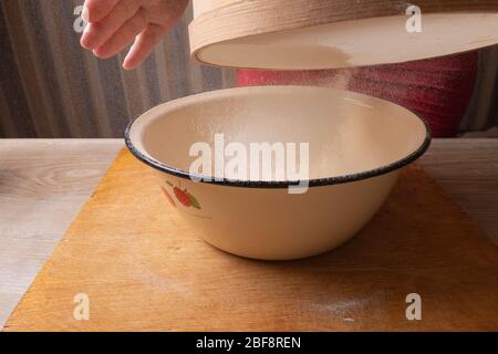 Große Schüssel, Schneidebrett, Sieb zum Sieben von Mehl auf dem Tisch. Weibliche Hände sieben Mehl in der Küche. Stockfoto