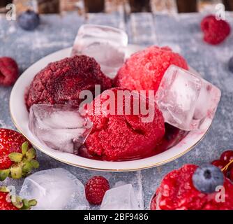 Kugeln aus selbstgemachter Erdbeere, roter Johannisbeere, Himbeere, Heidelbeereis, Sorbet in Schale, Kugeln, Beeren und Eiswürfel über auf blauem Holzbackgroun Stockfoto