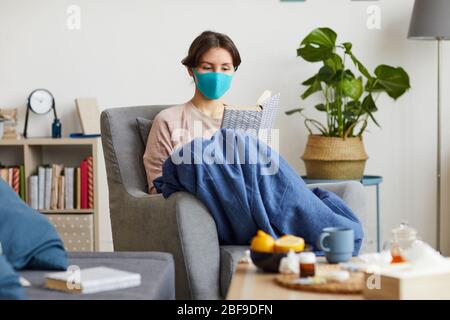 Junge kranke Frau in Schutzmaske sitzt auf dem Sessel und liest ein Buch sie zu Hause bleiben wegen der Epidemie Stockfoto