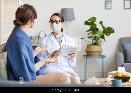 Junge Krankenschwester im weißen Mantel und in Schutzmaske, die den Tablet-pc hält und ihrem Patienten die Behandlungsmethode erklärt Stockfoto