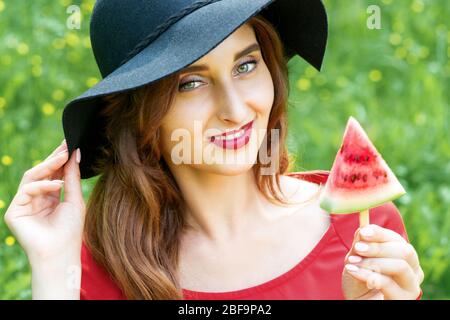 Modisch lächelnde junge Frau mit einem Stück Wassermelone in Form eines Dreiecks trägt schwarzen Hut auf grünem Hintergrund. Stockfoto