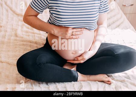 Schwangere Frau im T-Shirt hält die Hände auf dem Bauch auf einem weißen Hintergrund zu Hause, vor dem Fenster. Schwangerschaft, Mutterschaft, Vorbereitung und Erwartung werden übertrifft Stockfoto