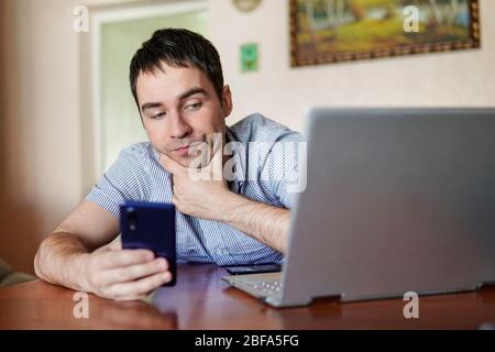 Geschäftsmann, der im Home Office mit einem Laptop arbeitet. Verbinden des Smartphones mit dem Computer über WLAN. Der Kerl arbeitet aus der Ferne vor dem Hintergrund von Stockfoto