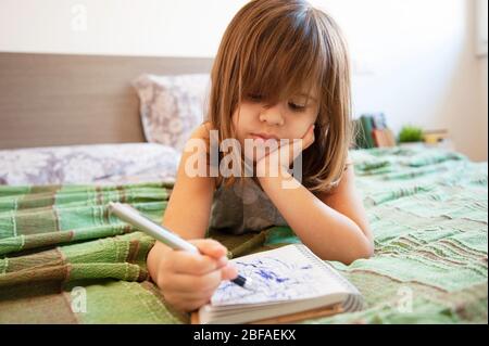 Kleine süße Vorschule Mädchen liegt auf dem Bett im Schlafzimmer zu Hause, sie ist gelangweilt und kritzeln mit blauen Stift auf dem Papier Notizbuch. Konzept der Kindheit. Lei Stockfoto