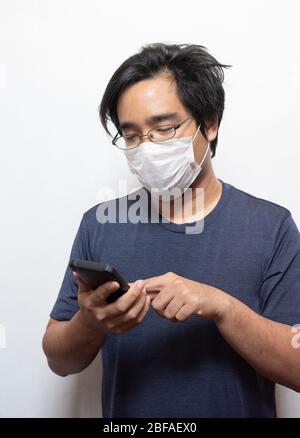 Asiatischer junger Mann in lässig tragen chirurgische medizinische Maske COVID-19 mit Smartphone auf weiß isoliert schützen Health Care Konzept.Wuhan coron Stockfoto