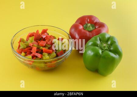 Rohe frische ganze rote und grüne Paprika und gekochte gehackte Paprika in Schüssel auf gelbem Hintergrund Stockfoto