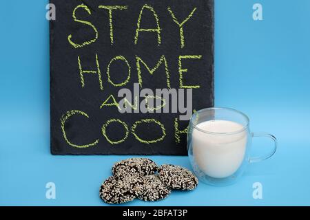 Ein Glas Milch und Schokoladenkekse auf blauem Hintergrund. Die Inschrift auf der schwarzen Tafel bleibt zu Hause und kocht. Das Konzept -Quarantäne, wh Stockfoto