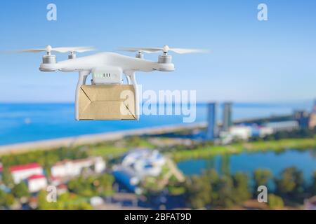 Technologische Sendung Innovation - Drohne schnelle Lieferung Konzept, Multicopter fliegen mit Karton über der Stadt in der Nähe von Meer Stockfoto