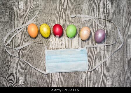 Viele farbige ostereier liegen in der Nähe der medizinischen Maske auf dem Holzhintergrund. Coronavirus Covid-19. Konzeptfoto. Ostern 2020. Stockfoto