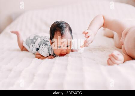 Ältere Bruder blonde junge Sohn mit kleinen Baby Schwester mit schwarzen Haaren auf dem Bett zu Hause Mädchen in grünen Körper kleiner Junge in Hosen spielen Stockfoto