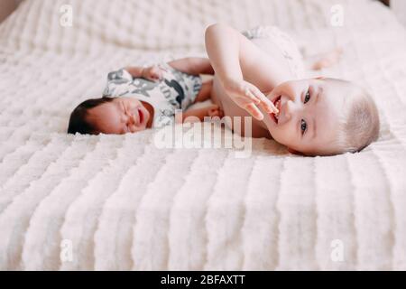 Ältere Bruder blonde junge Sohn mit kleinen Baby Schwester mit schwarzen Haaren auf dem Bett zu Hause Mädchen in grünen Körper kleiner Junge in Hosen spielen Stockfoto