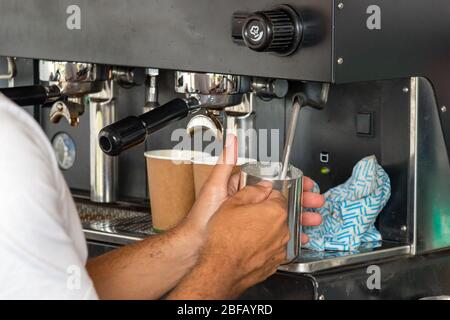 Der männliche Barista hält die Milch dampfend und bereitet einen Cappuccino zum Mitnehmen zu Stockfoto