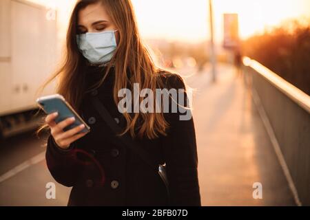 Junge Frau mit Schutzmaske Gesicht medizinische Maske mit Smartphone während der Spaziergang auf leeren Bürgersteig auf Brücke in der Stadt bei Sonnenuntergang Stockfoto