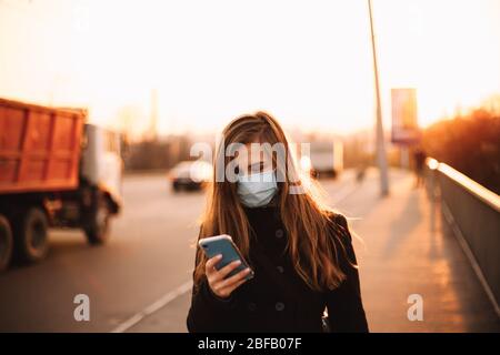 Glückliche junge Frau trägt Schutzmaske medizinische Smartphone beim Gehen auf leeren Bürgersteig auf Brücke in der Stadt bei Sonnenuntergang Stockfoto