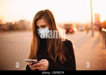 Ernste junge Frau trägt schützende Gesicht medizinische Maske mit Smartphone während des Spaziergangs auf leeren Bürgersteig in der Stadt bei Sonnenuntergang Stockfoto