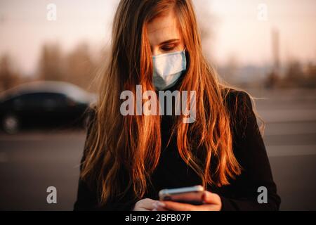 Porträt einer jungen Frau mit Schutzmaske Gesicht medizinische Maske mit Smartphone während auf der Straße in der Stadt bei Sonnenuntergang stehen Stockfoto