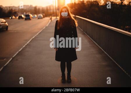 Porträt der jungen Frau mit Schutzmaske Gesicht medizinische Maske während auf leeren Bürgersteig auf Brücke in der Stadt bei Sonnenuntergang stehen