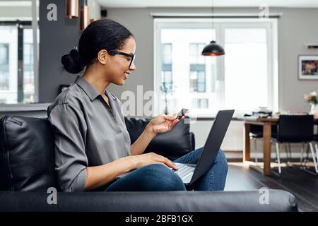 Seitenansicht der lächelnden Frau, die eine Karte mit Laptop auf den Hüften zu Hause hält Stockfoto