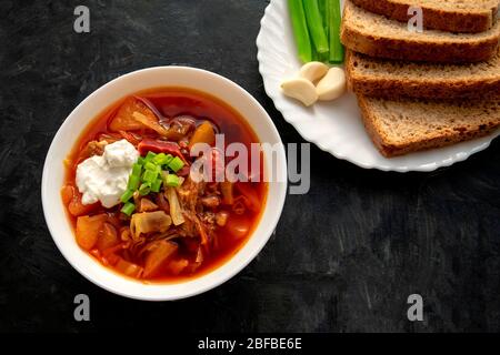 Borscht-Suppe in einer weißen Schüssel mit schwarzem Brot, grüner Zwiebel und Knoblauch auf dunkelgrauem Hintergrund. Berühmte traditionelle ukrainische und russische Borschtschensuppe w Stockfoto