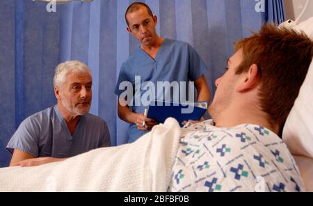 Ein bärtiger Chirurg und ein Mitglied seines Operationsteams besuchen einen männlichen Patienten, der sich nach einer Operation im Bett erholt Stockfoto