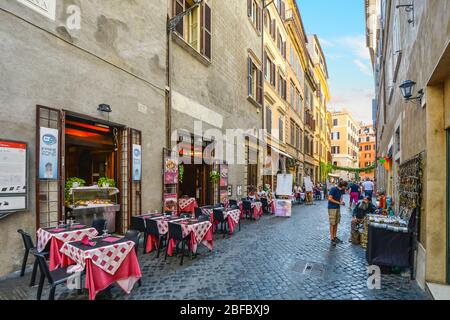 Buntes italienisches Restaurant mit Tischen in einem Straßencafé in einer Gasse in Rom, Italien, mit einem Straßenverkäufer, der Souvenirs an einen Kunden verkauft. Stockfoto