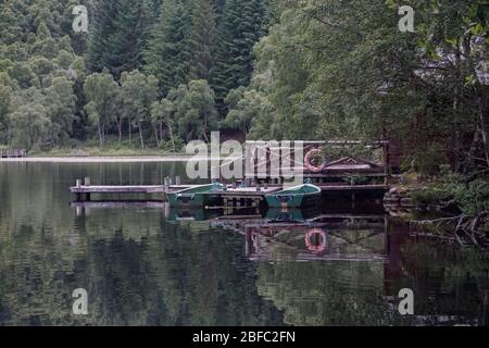 Ruderboote an einem Dock an einem Deck einer Hütte auf einem ruhigen See in den Highlands von Schottland gebunden. Stockfoto