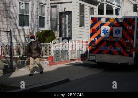 Chelsea, Massachusetts, USA. April 2020. Die COVID-19, Corona Virus, Situation in Chelsea, Massachusetts ist ein wichtiger Hotspot und die am härtesten getroffenen Stadt im Bundesstaat. Chelsea ist eine lateinamerikanische Einwandererbevölkerung mit 80% der Bevölkerung sind wichtige Arbeitskräfte, wie Lebensmittelarbeiter. Ein Mann geht die Straße hinunter in einer Maske an einem Krankenwagen vorbei. Die Ambulanzen arbeiten Tag und Nacht ununterbrochen mit COVID-Anrufen. Kredit: Allison Dinner/ZUMA Wire/Alamy Live News Stockfoto