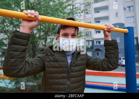 Teenager-Junge steht in der Nähe horizontale Bar von Outdoor-Trainingsgeräten, auf Spielplatz in der Nähe von Hochhäusern mit Wohnungen, eine medizinische Maske auf seinem Gesicht p Stockfoto