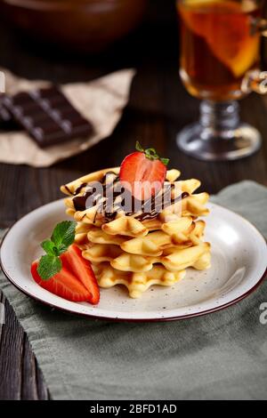 Wunderschönes Frühstück. Hausgemachte belgische Wiener Waffeln mit Schokolade und Erdbeeren, Tee auf dunklem Holzhintergrund dekoriert. Stockfoto