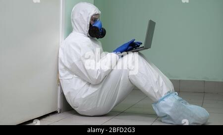 Mittlere Aufnahme. Müde Arzt in Schutzkleidung sitzt auf dem Boden gegen die Wand gelehnt arbeitet auf seinem Laptop. Professionelle Aufnahme in 4K-Auflösung. 054. Stockfoto