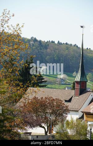 Altes reformiertes Kirchengebäude mit Kupfer- und Fliesendach in Urdorf, Schweiz unter Bäumen, Seitenansicht mit Dorfschild oben. Stockfoto