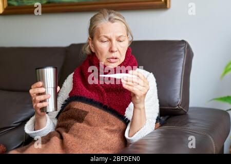 Die Frau hat eine Grippe und sie benutzt ein Thermometer. Krank mit einer Rhinitis Frau tropft Nase. Frau, die krank ist und Grippe hat, liegt auf dem Sofa und schaut tempe an Stockfoto