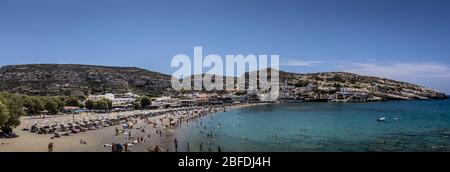 Die Küste und Berge von Matala an einem klaren Sommer sonnigen Tag mit und einem blauen Himmel im Hintergrund. Kreta, Griechenland. Sonnenliegen am Strand mit Tour Stockfoto