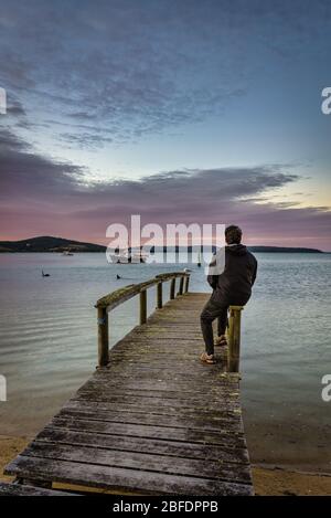 Touristen sitzen auf einem alten Holzsteg und sehen den perfekten Sonnenuntergang am Hafen, mit Booten, die sicher in St Helen''s Bay in Tasmanien verankert sind. Stockfoto