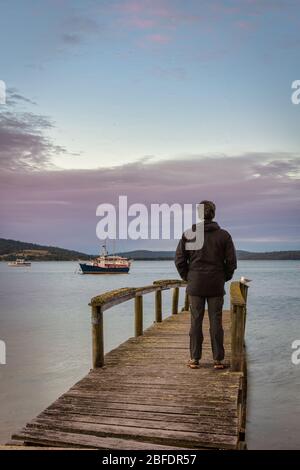 Touristen stehen auf einem alten hölzernen Steg und sehen den perfekten Sonnenuntergang am Hafen, mit Booten sicher in St Helen''s Bay in Tasmanien verankert. Stockfoto