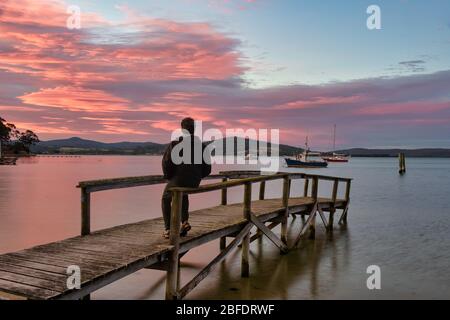 Touristen sitzen auf einem alten Holzsteg und sehen den perfekten Sonnenuntergang am Hafen, mit Booten, die sicher in St Helen''s Bay in Tasmanien verankert sind. Stockfoto