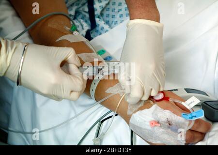 Die Hand eines Krankenpflegers mit Handschuhen injiziert Flüssigkeit in einen Katheter, der am Arm eines Patienten befestigt ist. Stockfoto