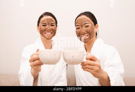 Zwei Freunde mit Schlammmasken auf ihren Gesichtern trinken Tee. Spa-Entspannung, Gesichtshautpflege, Hautreinigung und -Behandlung. Teil einer Serie mit Schlammmaske Stockfoto