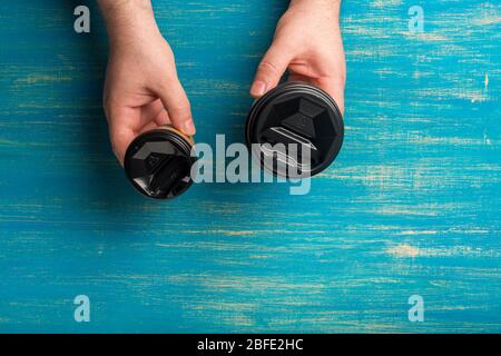 Zwei Einwegbecher aus Papier für Kaffee in einer männlichen Hand auf blauem Holzhintergrund. Stockfoto