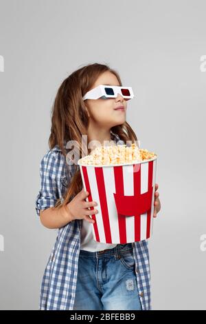 Nettes Mädchen, das rot-blaue 3d-Brille trägt und Popcorn aus Eimer auf grauem Hintergrund isoliert isst. Stockfoto