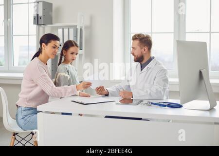 Der männliche Arzt gibt Mutter und Tochter ein Rezept, während er an einem Tisch in einem Klinikbüro sitzt. Stockfoto