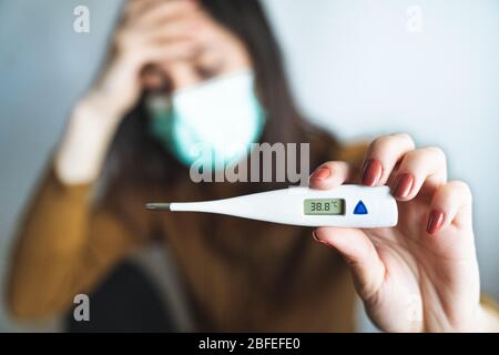 Selektiver Fokus einer kranken jungen Frau, die eine chirurgische Maske trägt und ein digitales Thermometer hält, das auf über 38 Grad Fieber hinweist. Krank und Stockfoto