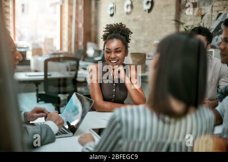 Wir sind ein Team! Junge und fröhliche Afro-amerikanerin lächelnd, während sie ein Treffen mit Kollegen im modernen Büro hat. Geschäftskonzept Stockfoto