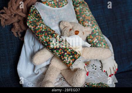 Eine handgemachte altmodische weibliche Puppe, bekleidet mit einem Kleid mit Schürzenkleid, sitzt auf einem blauen Sofa.Sie hält einen kleinen Teddybären in ihren Armen, er trägt ihn Stockfoto