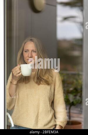 Blonde Frau im Pullover, die durch das Fenster schaut, Tee schlürft Stockfoto