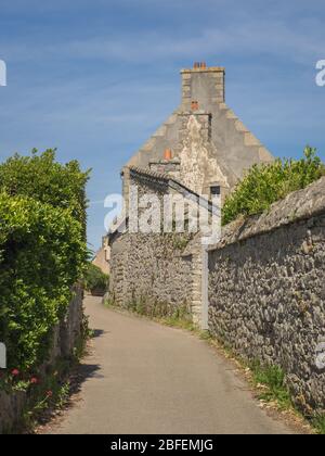 Steinmauern säumen eine schmale, leere Straße, die an einem sonnigen Sommertag in ein Dorf mit hoch aufragenden Schornsteinen in der Bretagne, Frankreich, führt. Stockfoto